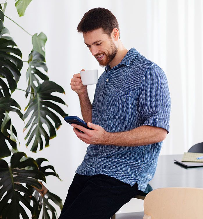 Smiling man leans on a kitchen island, sipping coffee while checking his phone in a bright home.